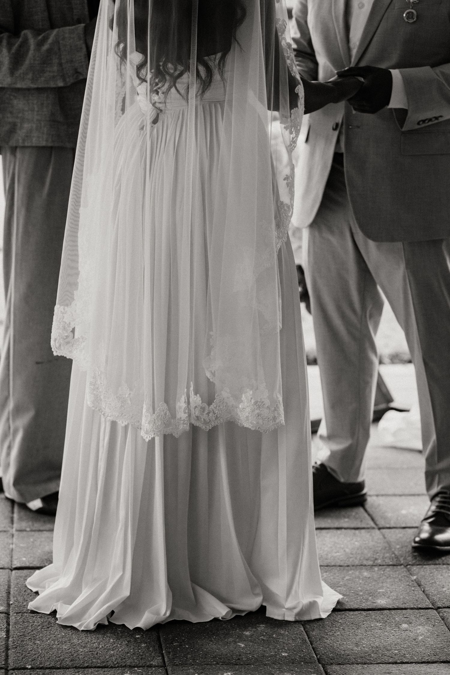Close-up detail of an elegant lace wedding dress and suit on stone pavement.