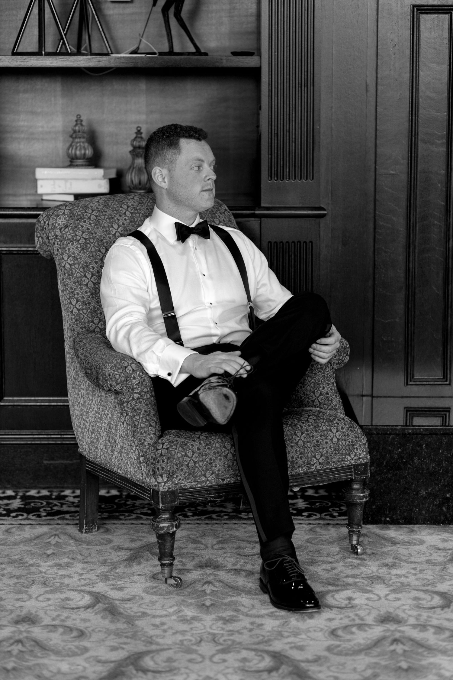 Black and white photo of someone seated in a vintage armchair wearing formal attire with suspenders.