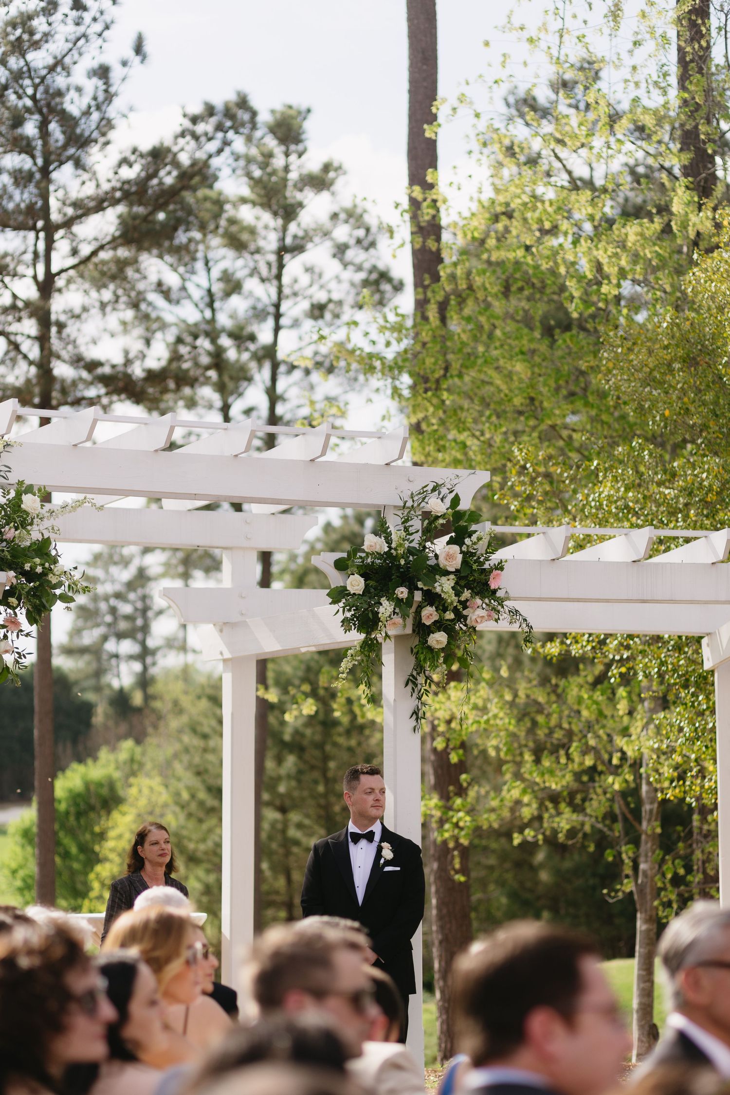 An outdoor wedding ceremony under a white pergola surrounded by pine trees.