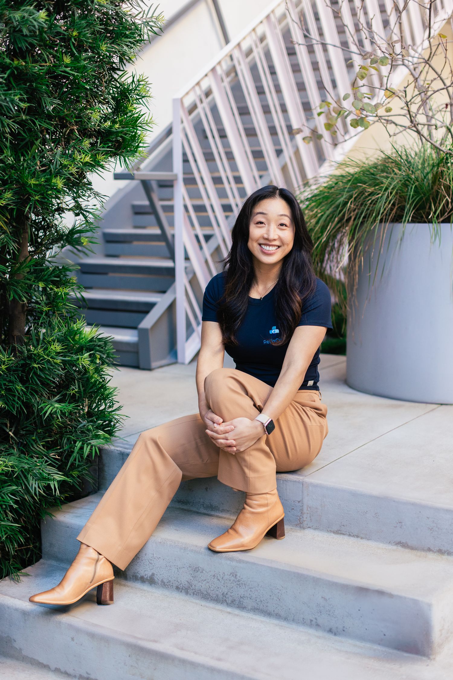 Someone sits smiling on outdoor concrete steps wearing a navy shirt, tan pants and brown boots near green plants and planters.