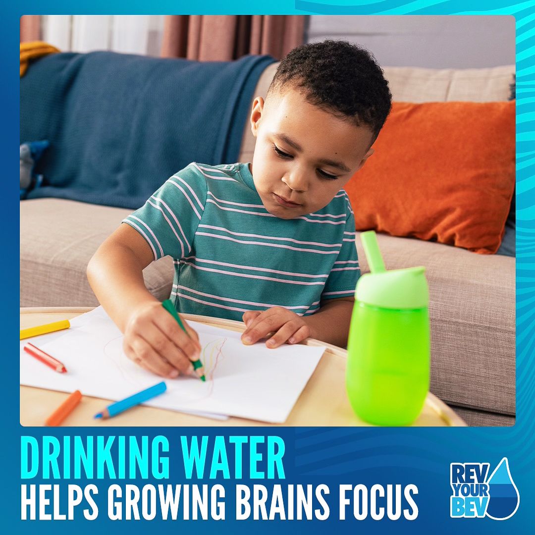 Young student in striped shirt sitting at desk while writing and drinking from green water bottle.