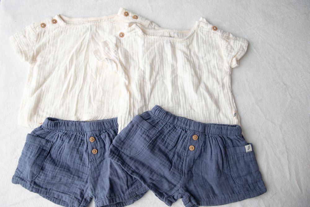 Two matching white muslin tops with wooden buttons paired with blue linen shorts for children laid flat.