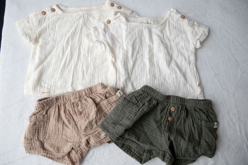 Two white muslin baby tops with wooden buttons displayed with pink and olive green linen shorts.