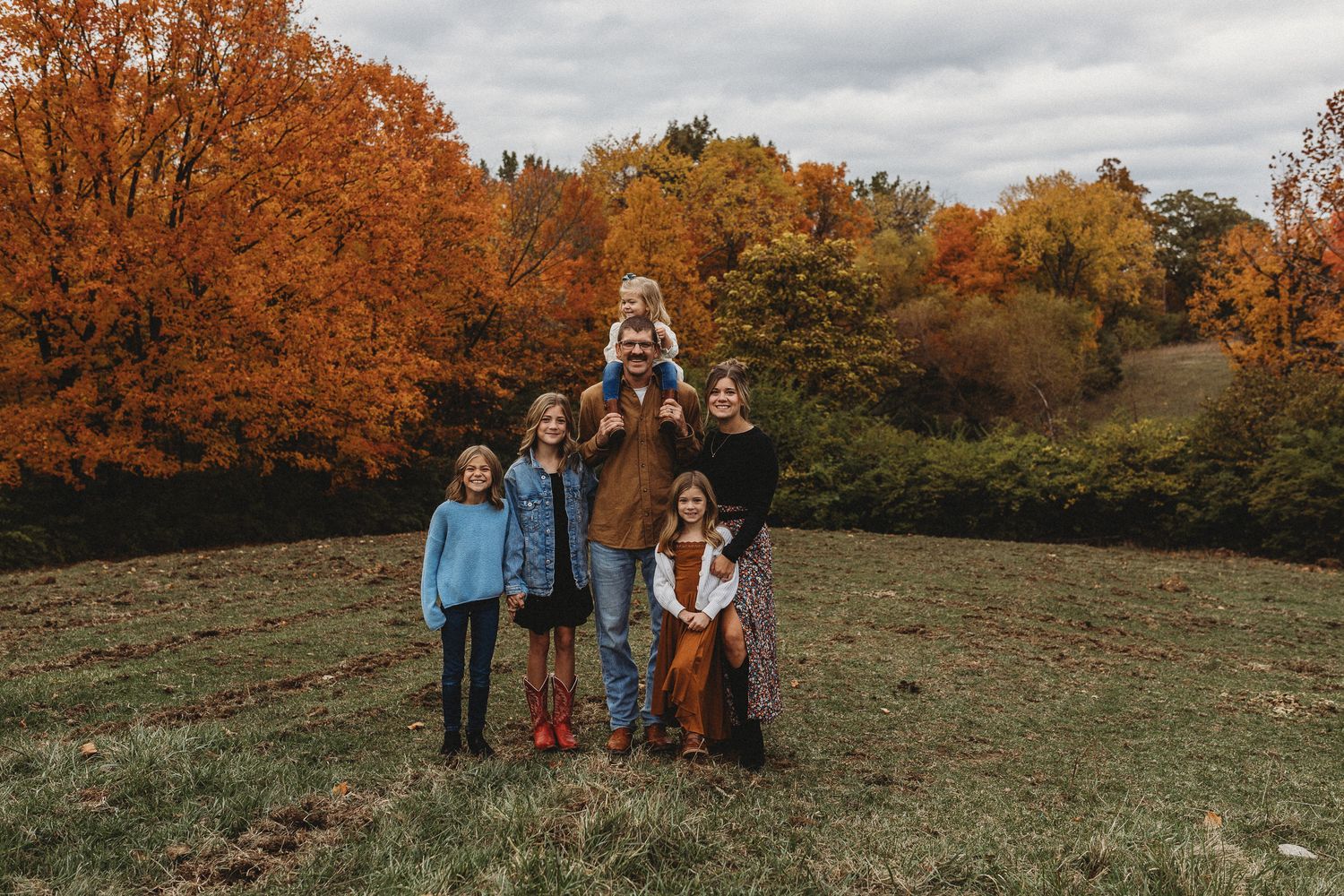 A group stands together in a grassy field surrounded by vibrant orange and red autumn trees on an overcast day.