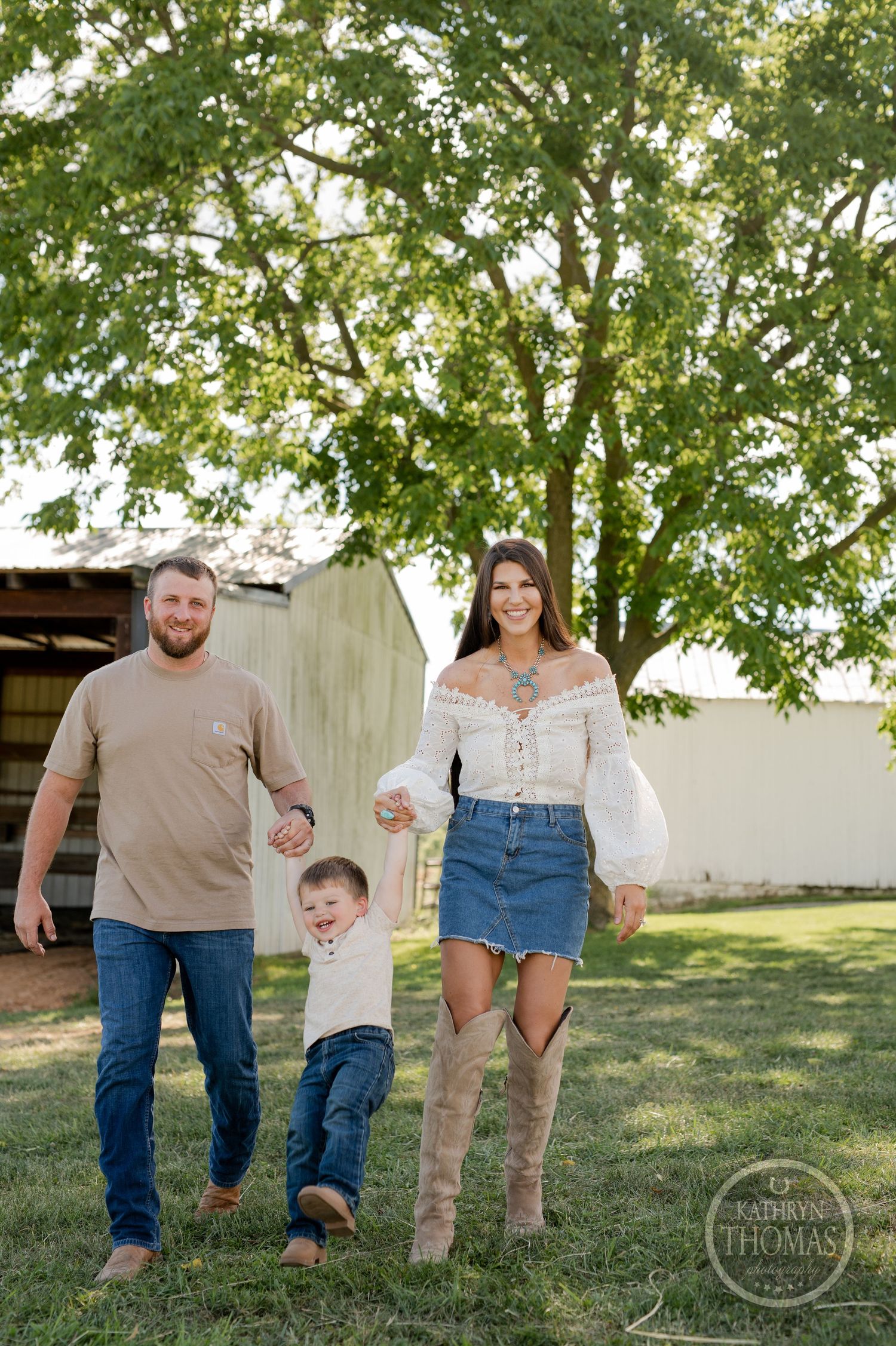 Family walking together outdoors holding hands near white barn under large tree.
