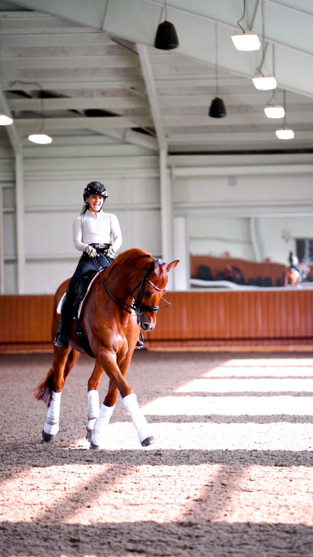 A rider performs dressage movements on a chestnut horse in an indoor arena with sunlight streaming through windows.