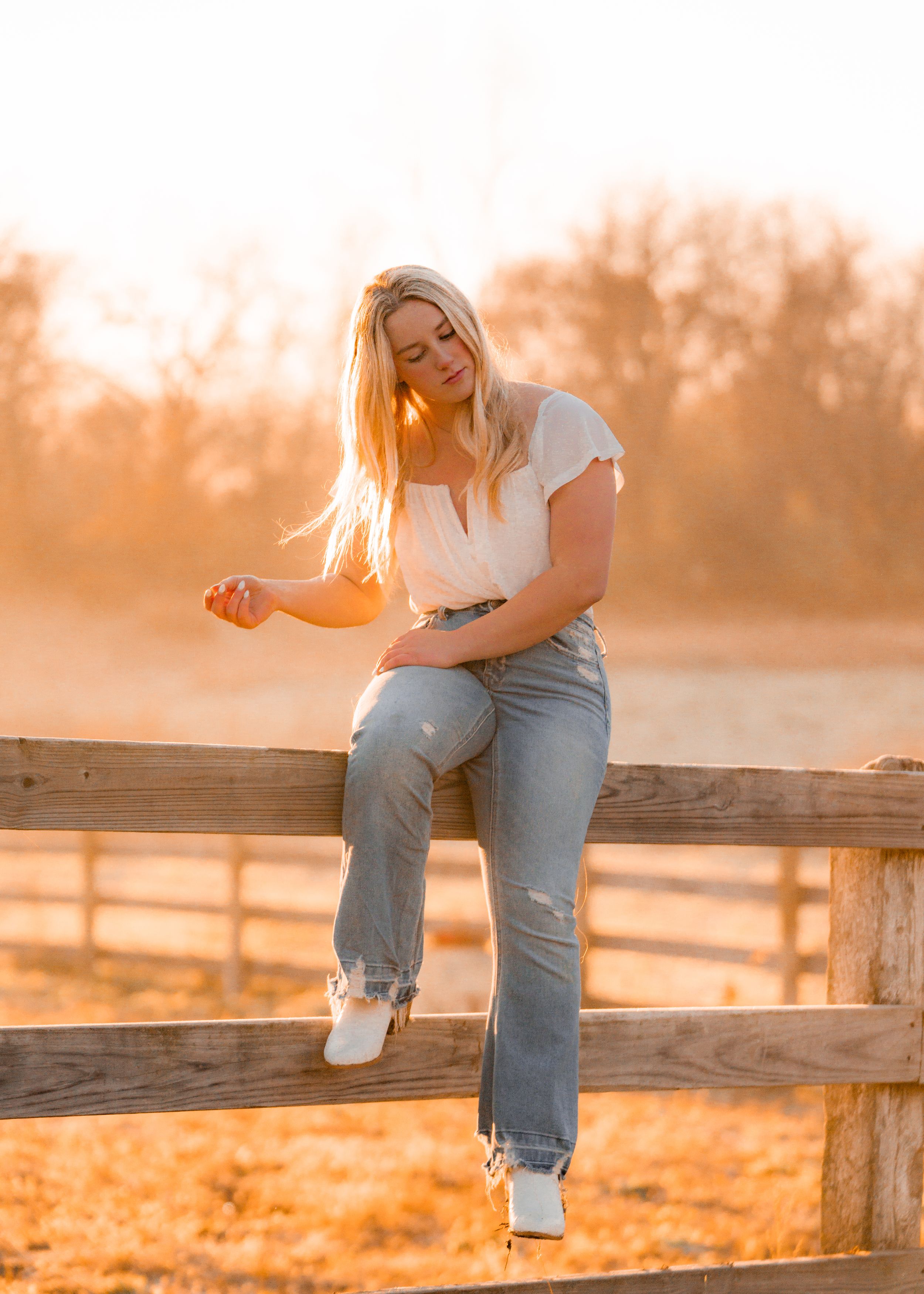 Person sitting on wooden fence during golden sunset wearing jeans and white top.