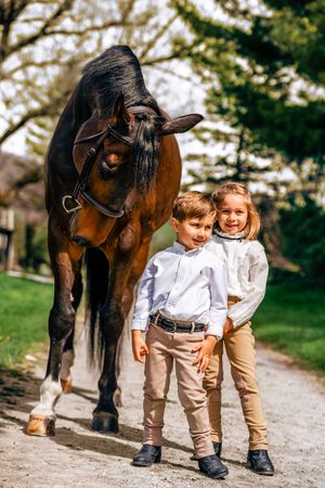 Two children stand beside a dark brown horse on a gravel path in a scenic outdoor setting.