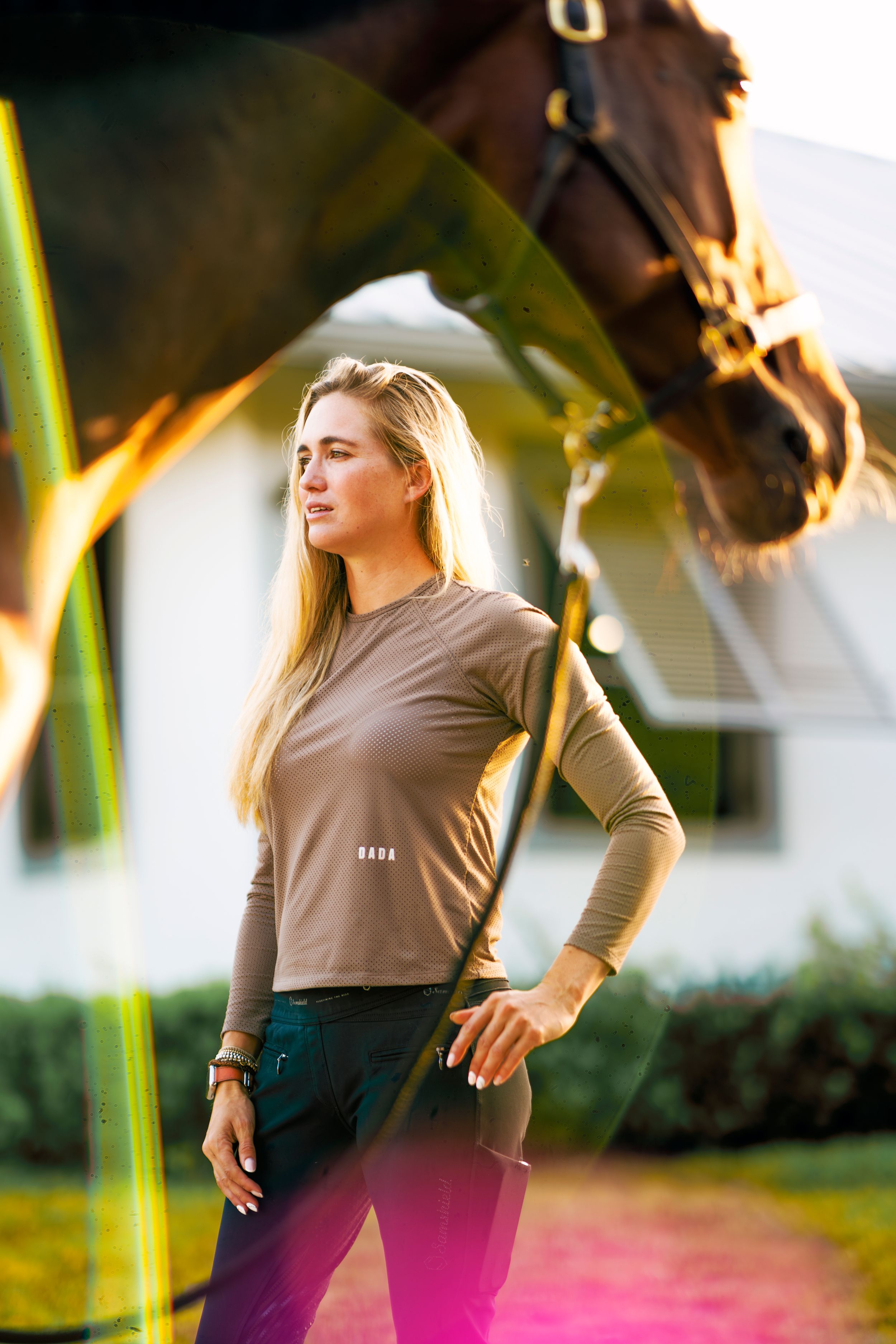 Woman in brown long sleeve top and dark pants poses with horse in outdoor setting at golden hour.