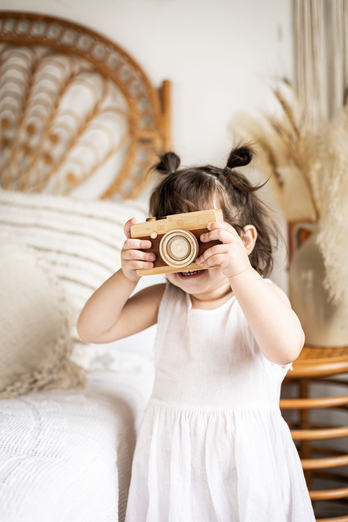 Small child in white dress holding a wooden toy camera while standing in a cozy boho-style room.