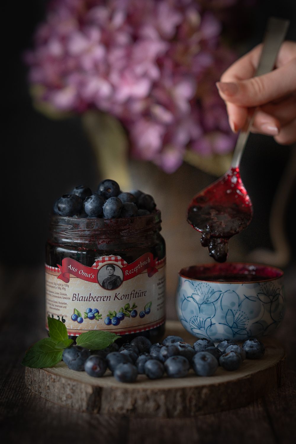 Foodfotografie. Fresh blueberries and jam on rustic wood with purple flowers in background.