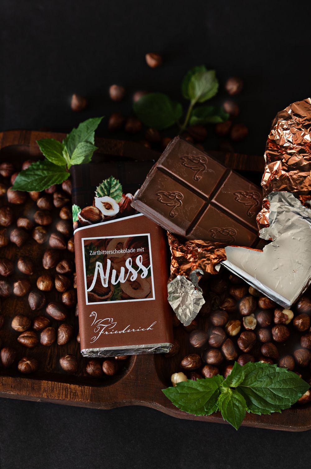 Foodfotografie. Dark chocolate bar with nuts scattered among coffee beans and mint leaves on wooden board.