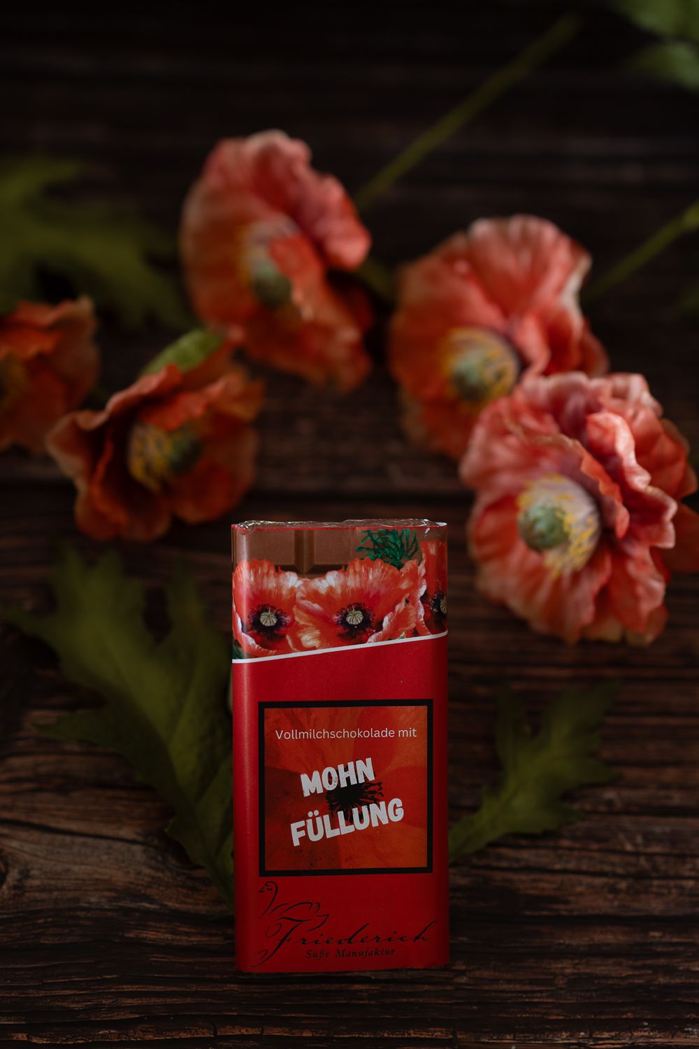 Foodfotografie. Red poppies arranged with a red bar of chocolate on rustic wooden surface featuring nostalgic floral styling.