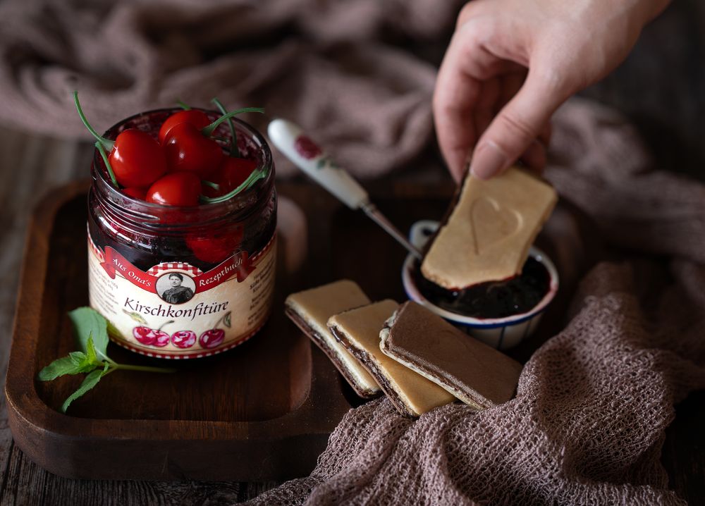 Food Fotografie - Glass jar of preserved cherries with biscuit and a hand holding one of them.