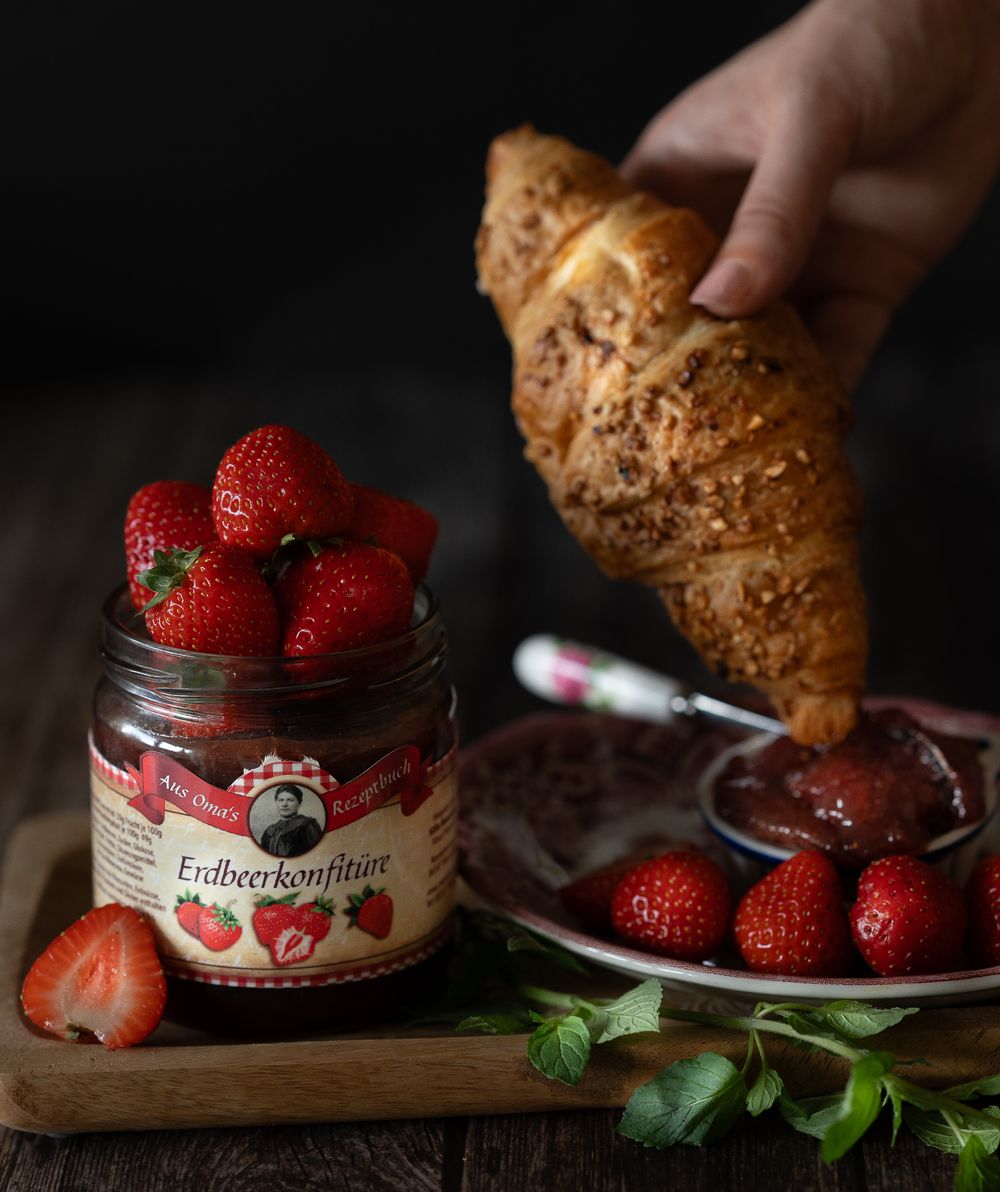 Foodfotografie. Fresh strawberries and jam jar beside croissant on dark wooden surface.