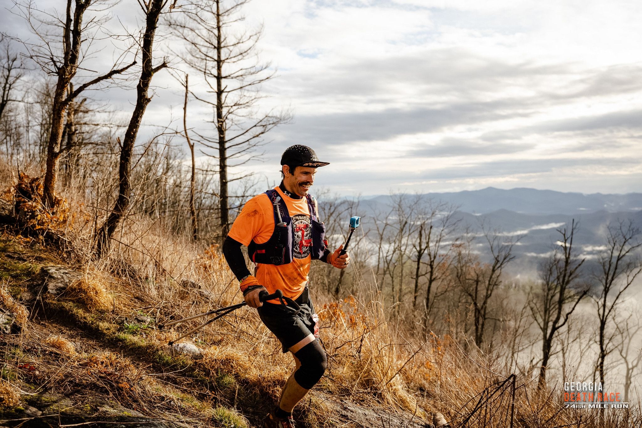 Trail runner in orange vest traverses mountain ridge with scenic valley views below.