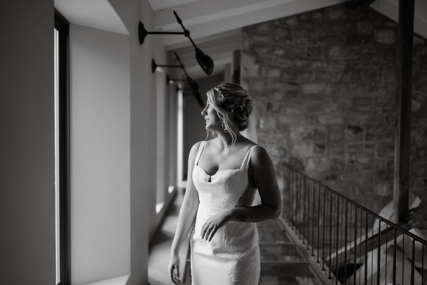 A bride in a fitted white dress stands near a brick wall in dramatic lighting.