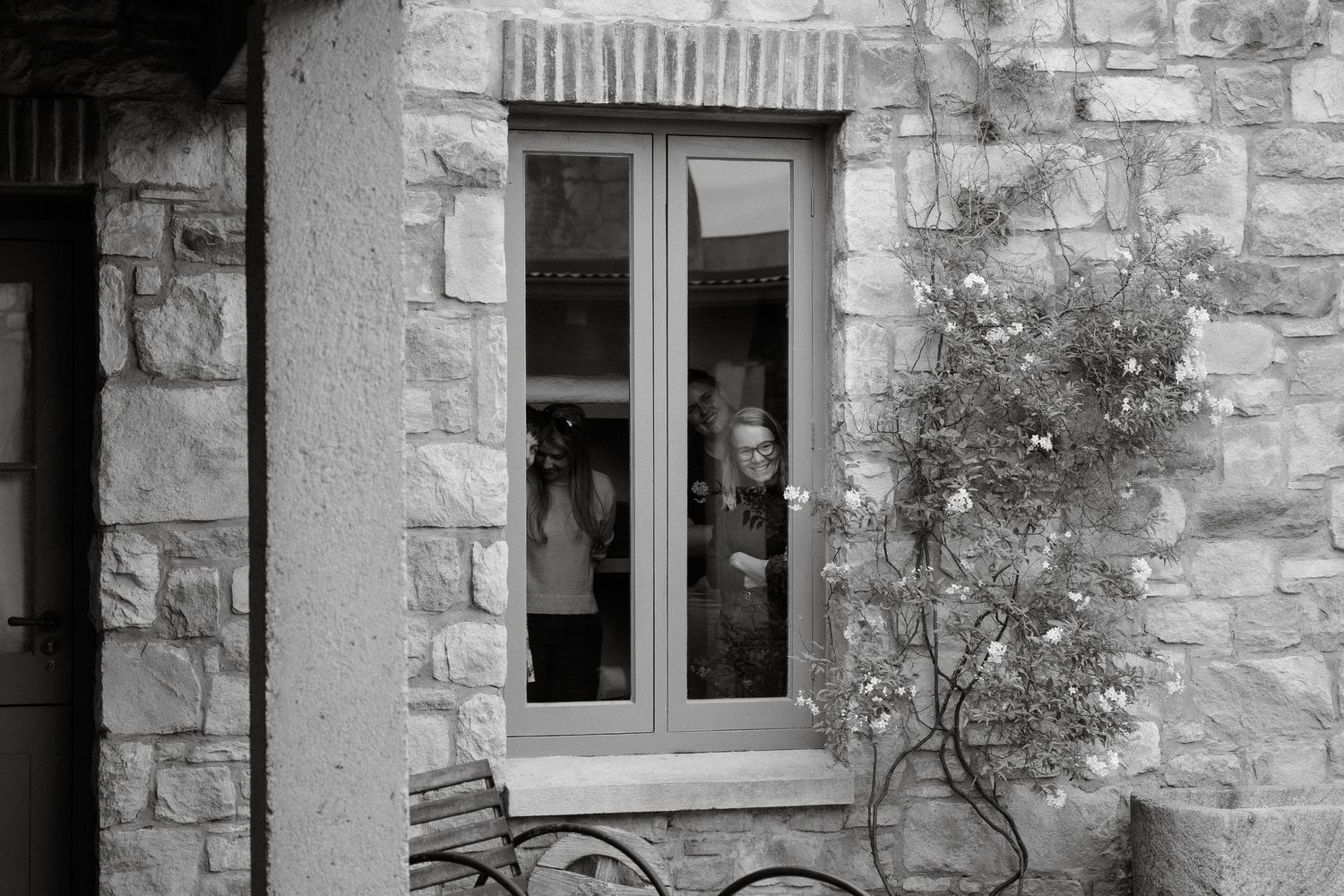 Black and white view through a stone window frame with someone peering out from inside a rustic building.