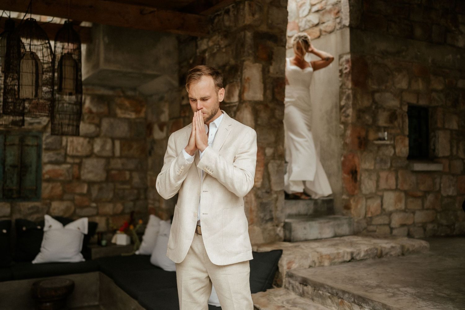A person in a light-colored suit stands thoughtfully against a rustic stone wall backdrop.
