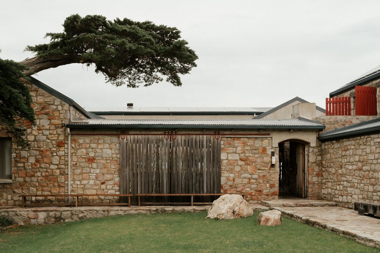 A modern rustic stone house with wooden door features and a large overhanging tree against an overcast sky.