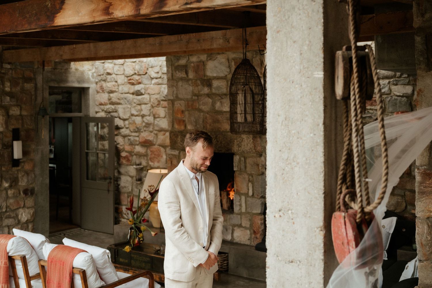 A person in a cream-colored suit stands in front of a rustic stone wall with wooden beams overhead.