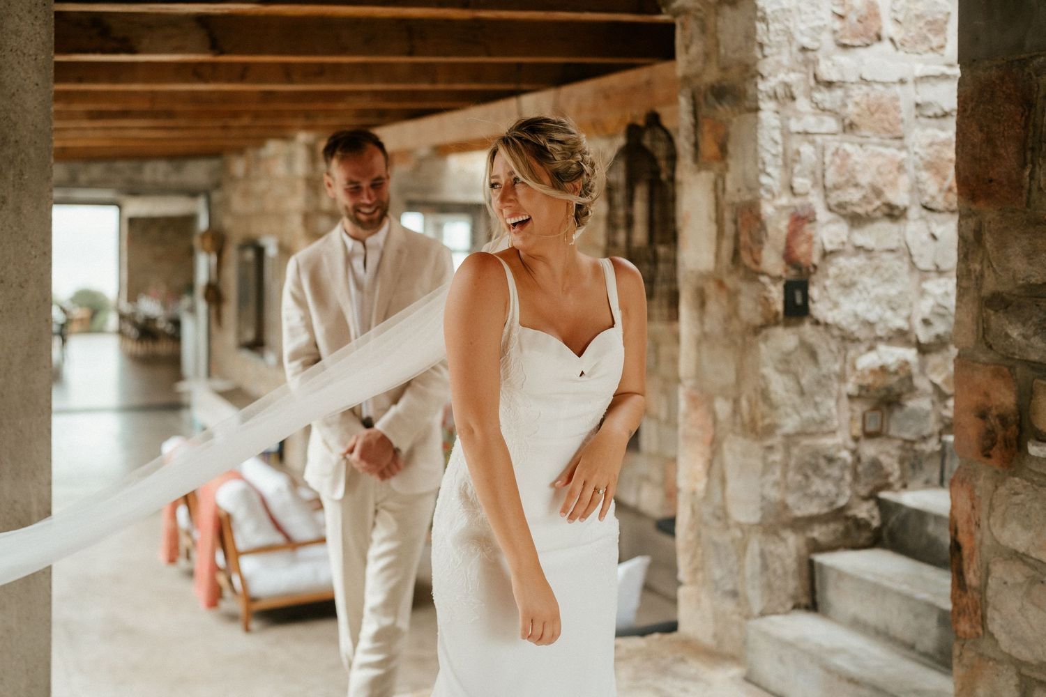 A couple in elegant white wedding attire poses playfully in a rustic stone building with natural light streaming in.