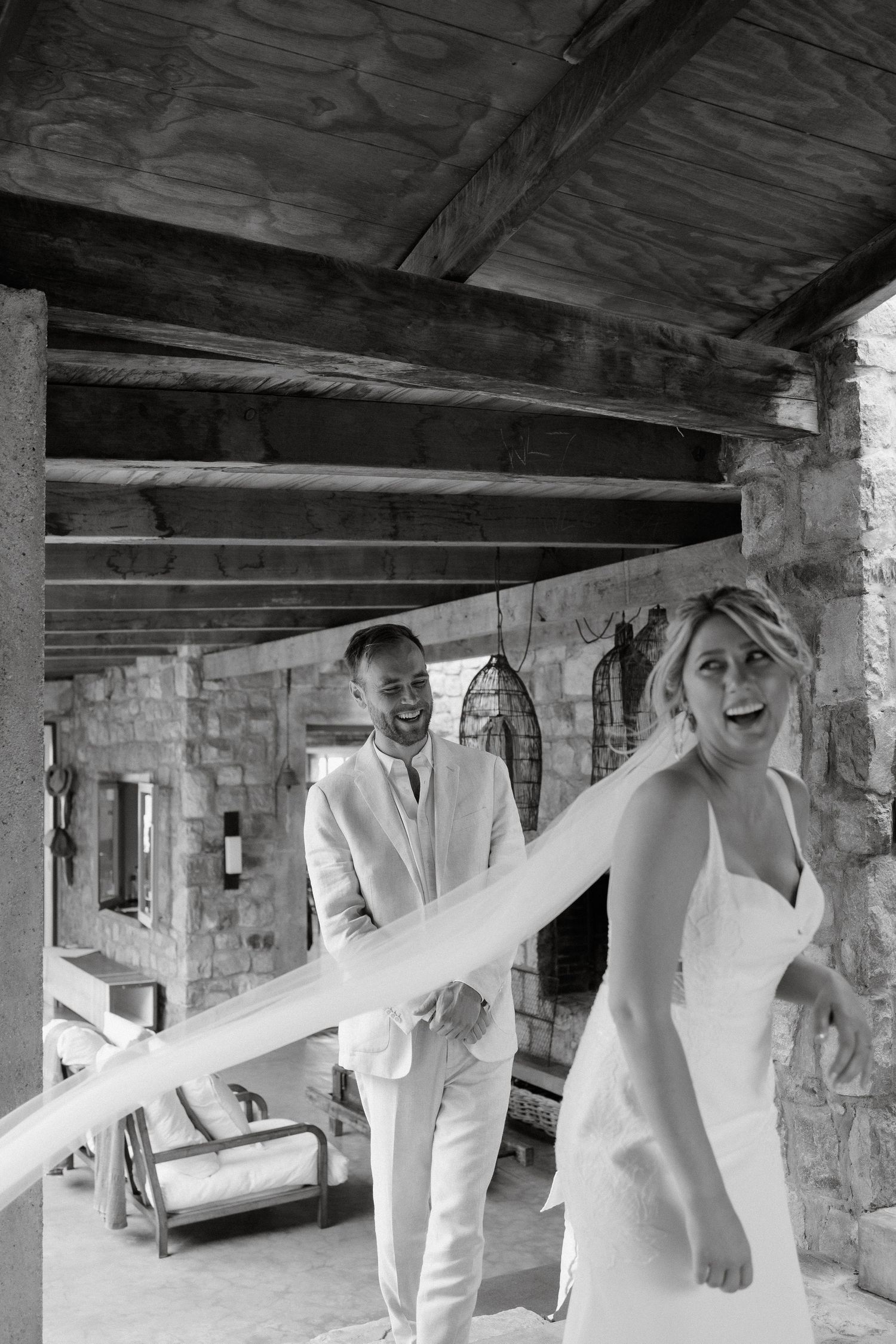 A joyful wedding couple in white attire laughing together under a rustic stone portico in black and white.