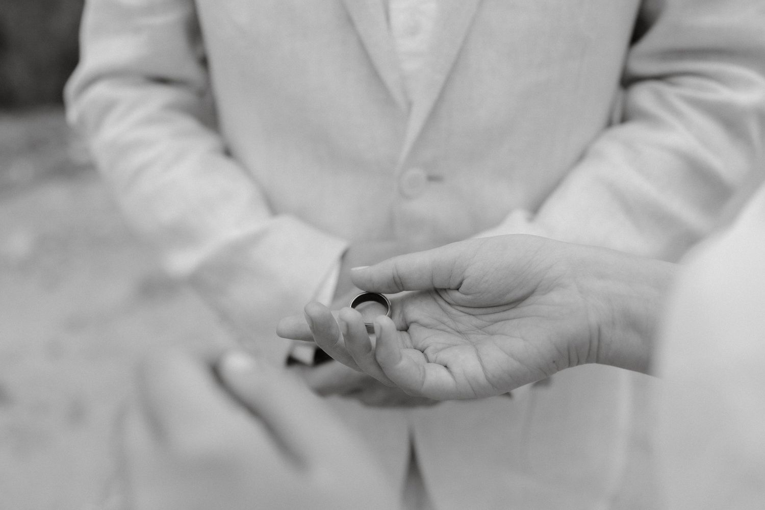 A black and white close-up photo shows wedding ring exchange between two hands during a ceremony.