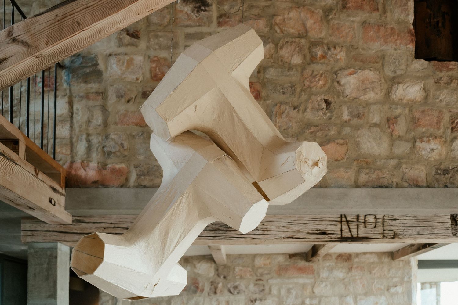A sculptural white dress form hangs suspended from rustic wooden beams against an exposed brick wall.