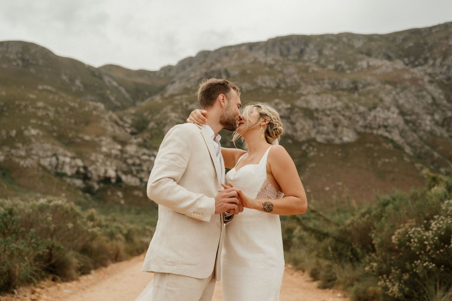 A joyful wedding couple in matching cream outfits laughing together on a mountain path.