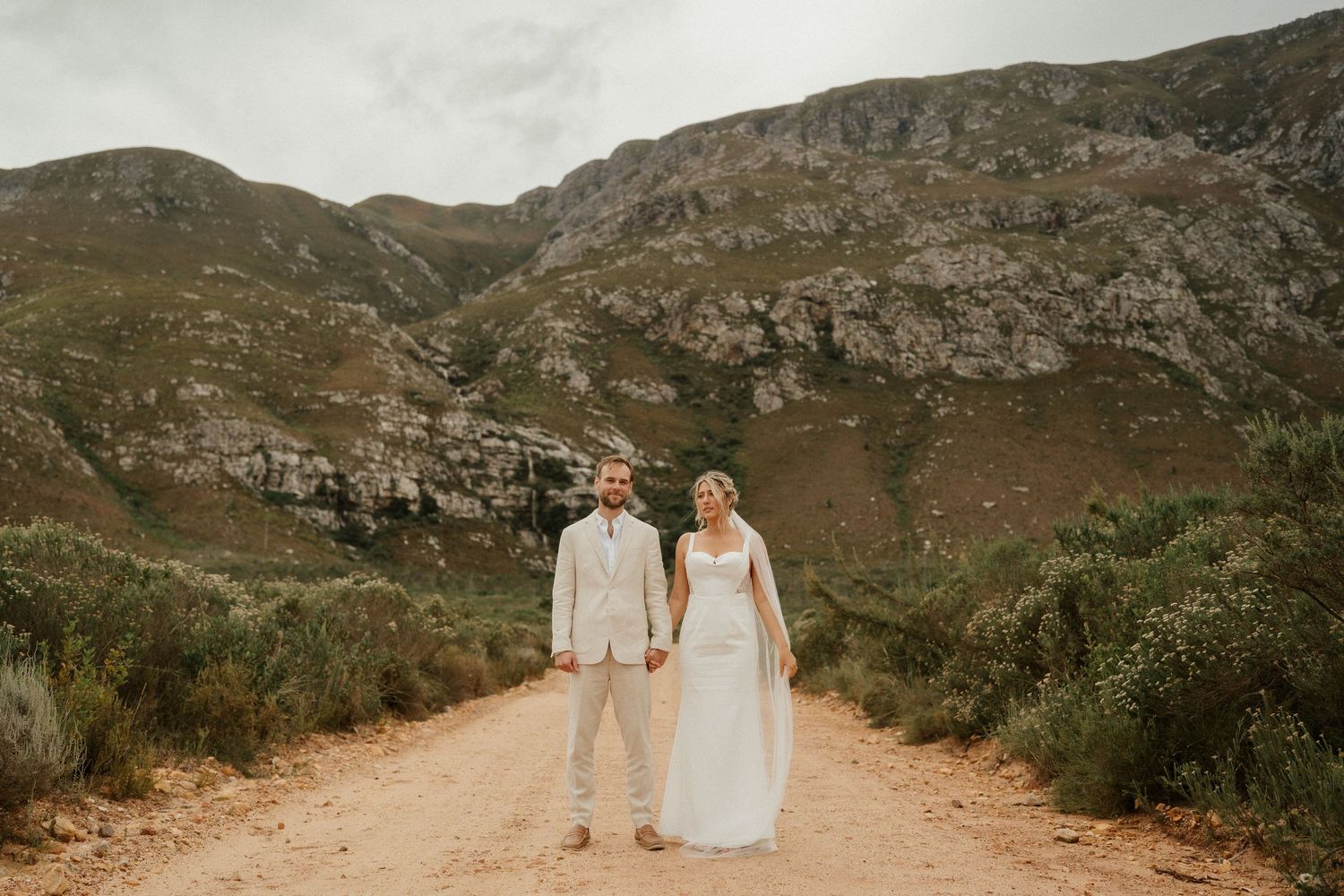 A couple stands on a dirt path surrounded by dramatic mountain landscapes on their wedding day.