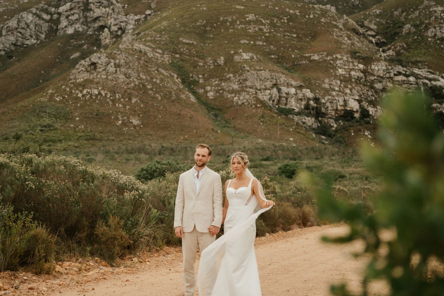 A bride and groom in light-colored attire pose together on a dirt path with rugged mountains behind them.