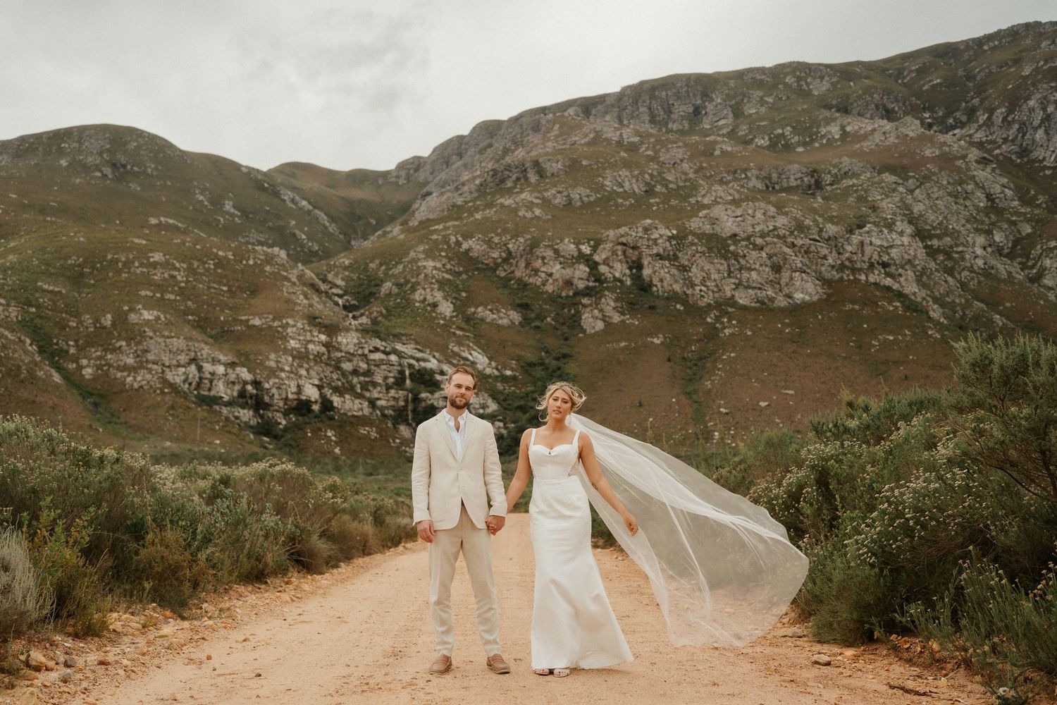 A couple in wedding attire stands embracing on a dirt path with dramatic mountain scenery in the background.