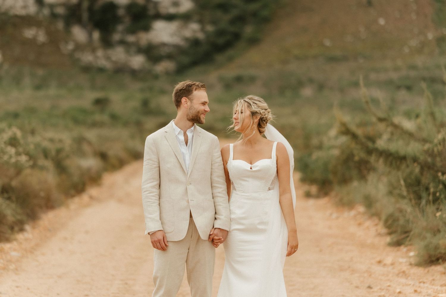 A couple in wedding attire walks hand in hand down a scenic dirt path with mountains in the background.
