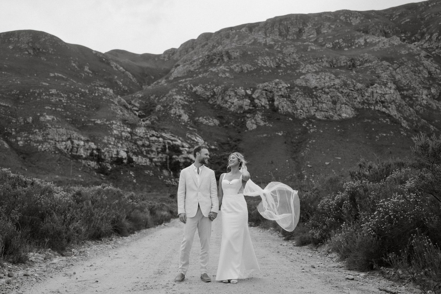 A romantic couple in wedding attire poses on a dirt path with dramatic mountain scenery in black and white.