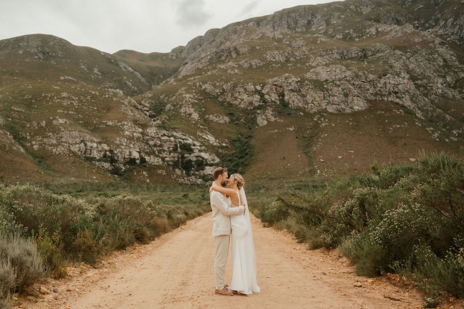 Newlyweds embrace on a dusty mountain road with dramatic rocky cliffs behind them.