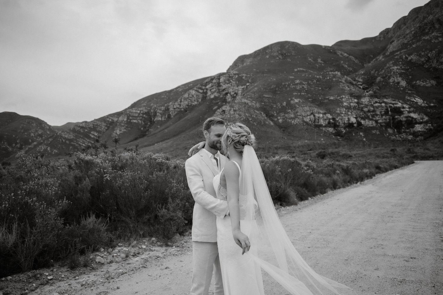 A romantic wedding couple embraces on a dirt road with dramatic mountainous landscape in black and white.
