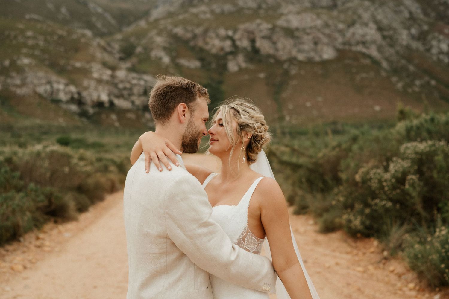 A romantic wedding moment captured on a dirt path with rugged mountains in the background.