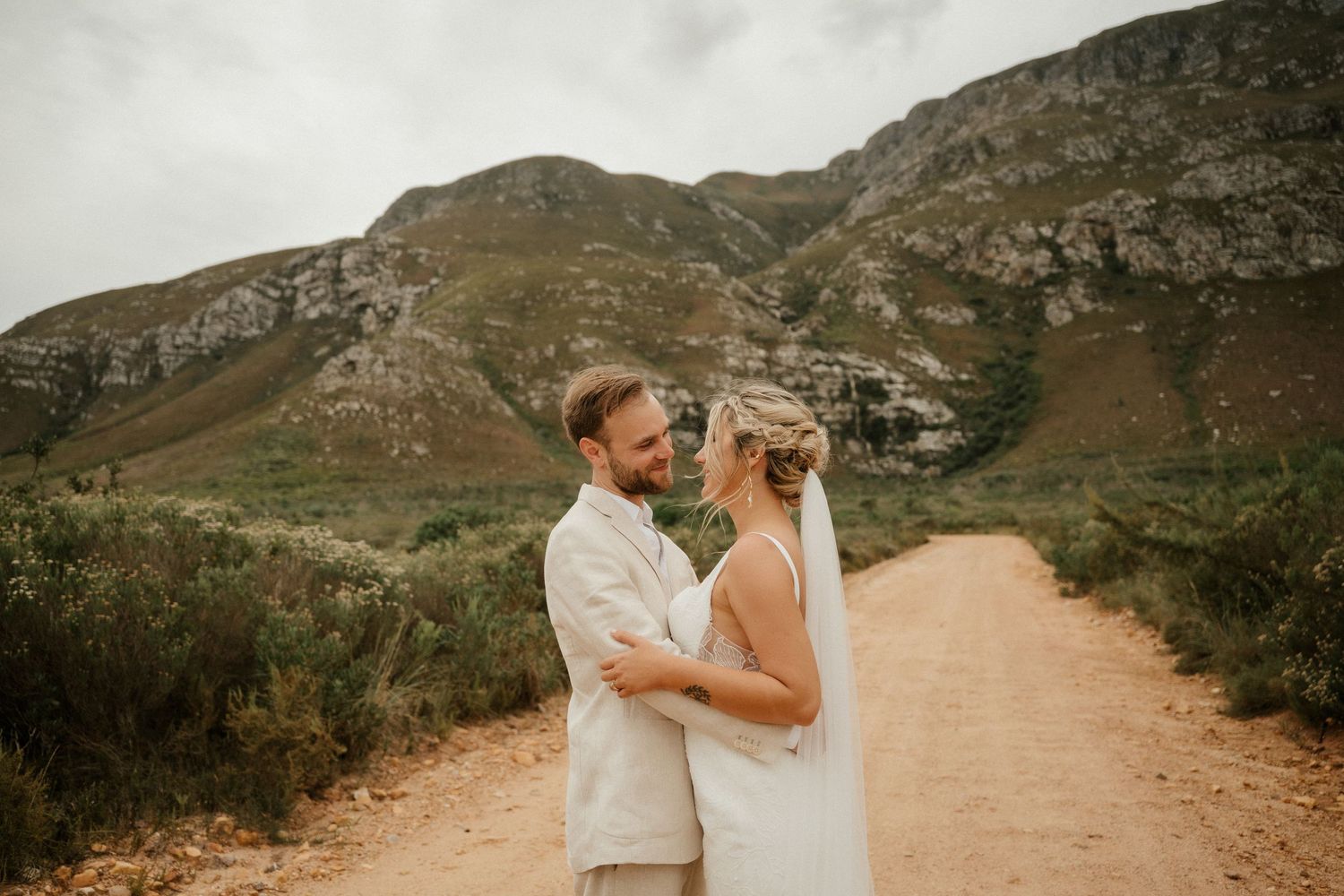 A wedding couple embraces on a dirt road with dramatic mountain cliffs rising in the background.