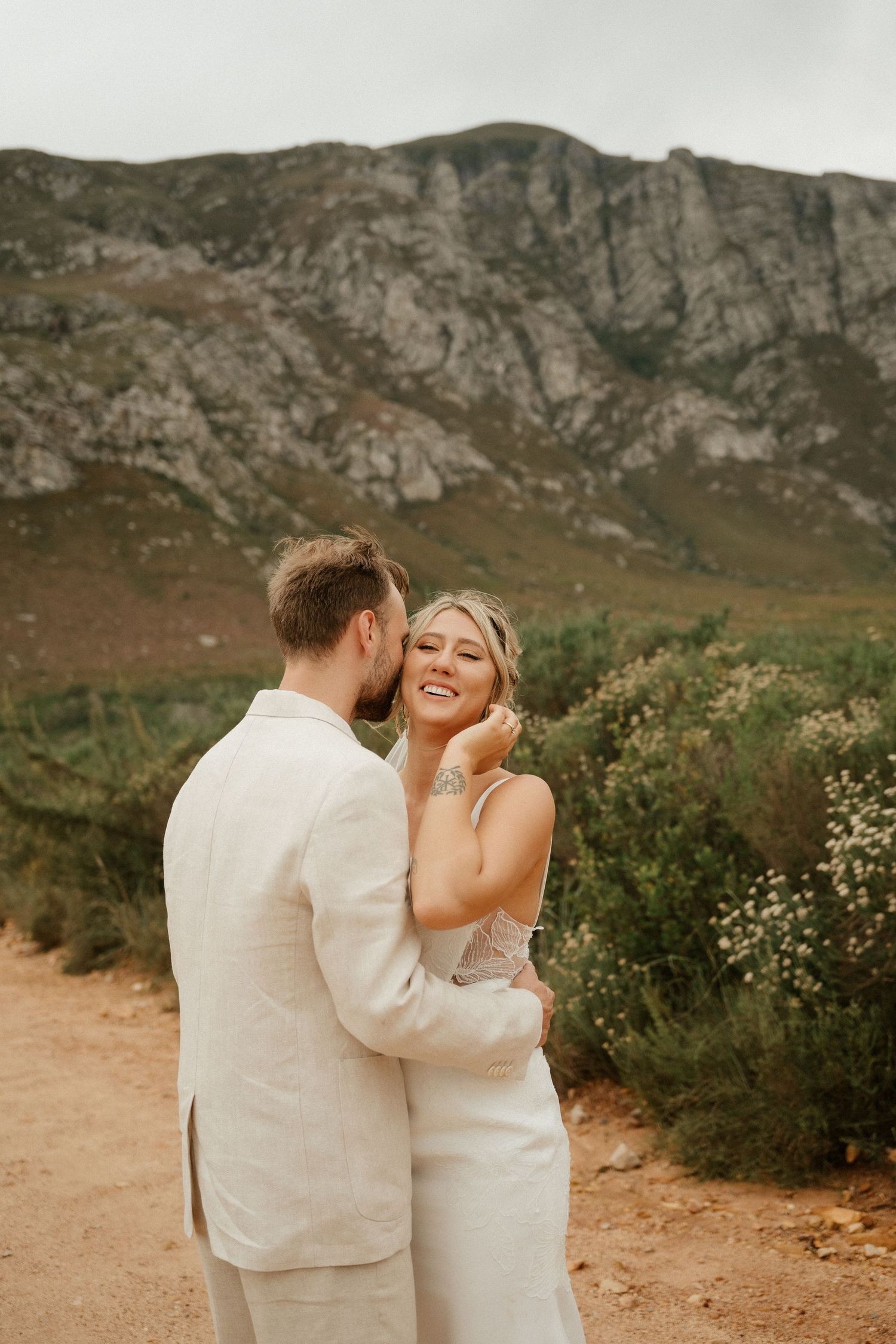 Newlyweds share an intimate moment on a desert path with rugged mountains towering behind them.