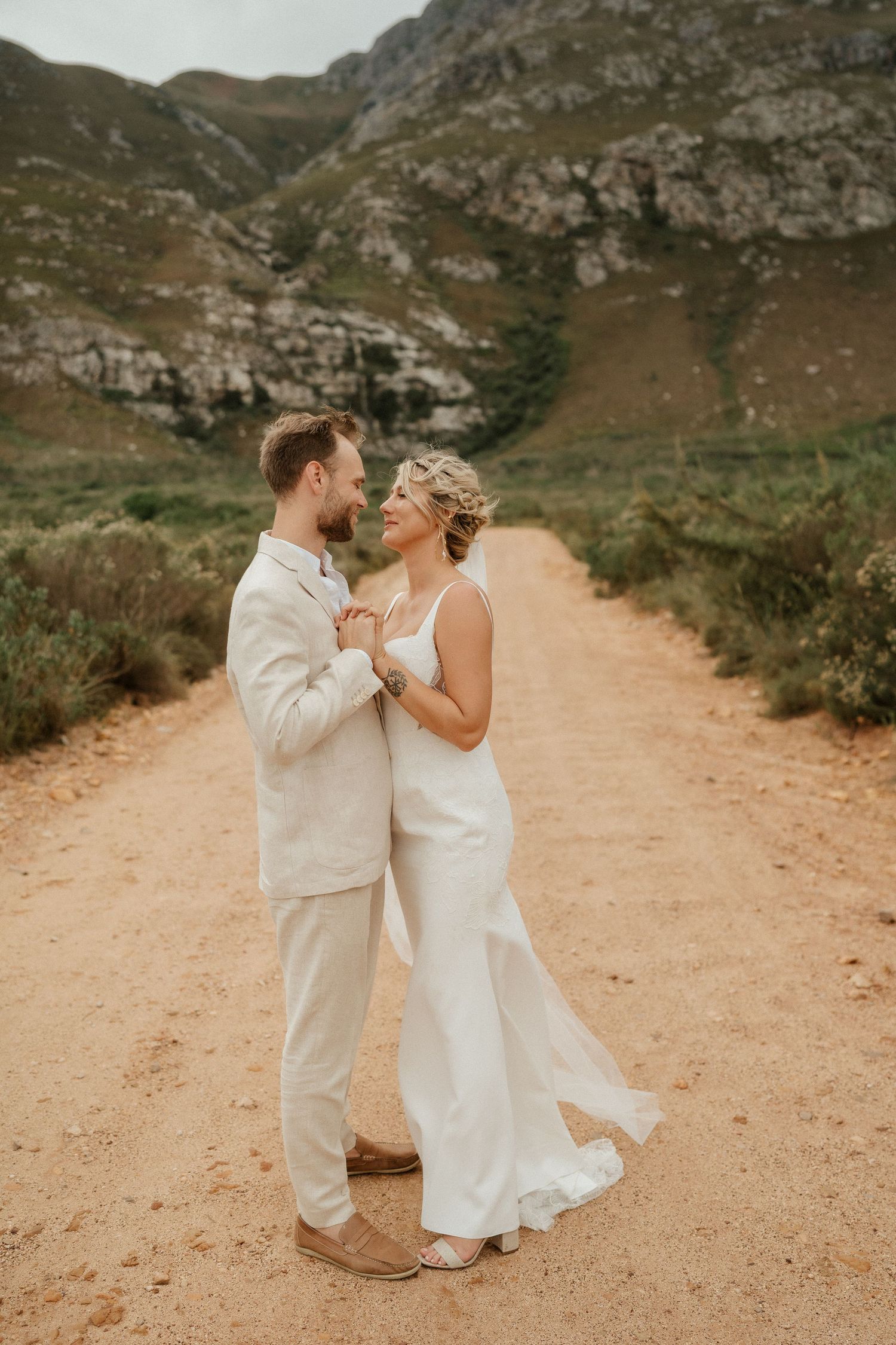 A couple in white wedding attire dance together on a mountain path during their special day.
