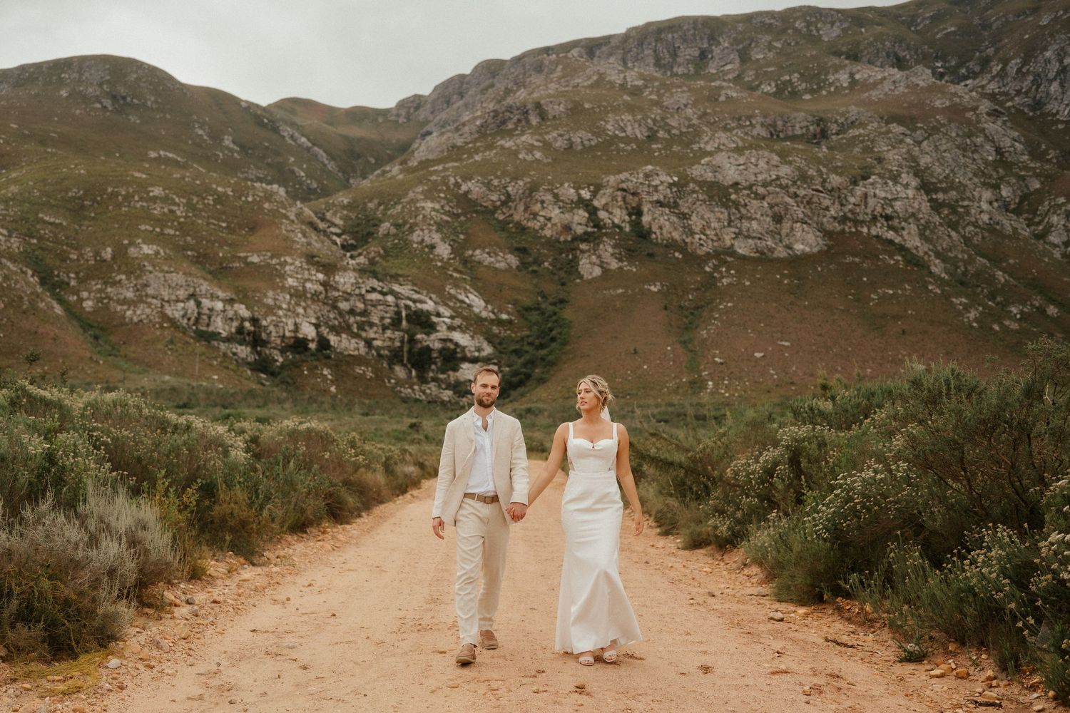 A couple in wedding attire walking hand in hand down a scenic mountain dirt road.