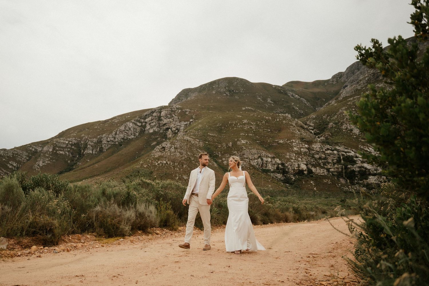 Newlyweds in white walk hand-in-hand along a scenic mountain trail during their intimate wedding.