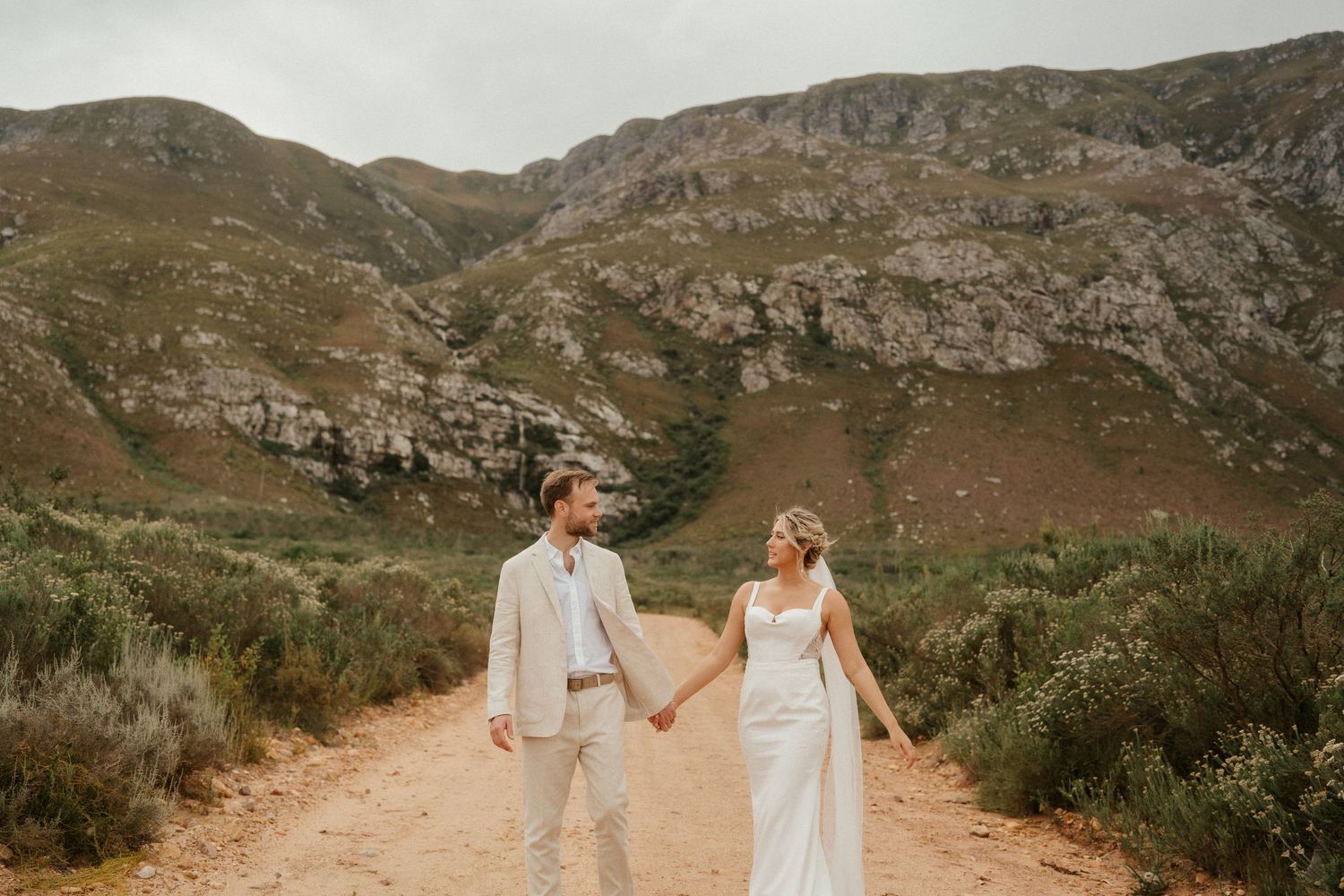 A couple in wedding attire walks hand in hand on a dirt path with rugged mountains in the background.
