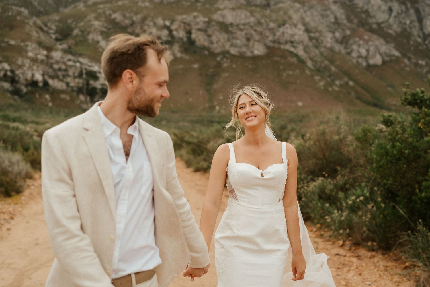 Wedding couple in white attire share a joyful moment on a mountain trail.