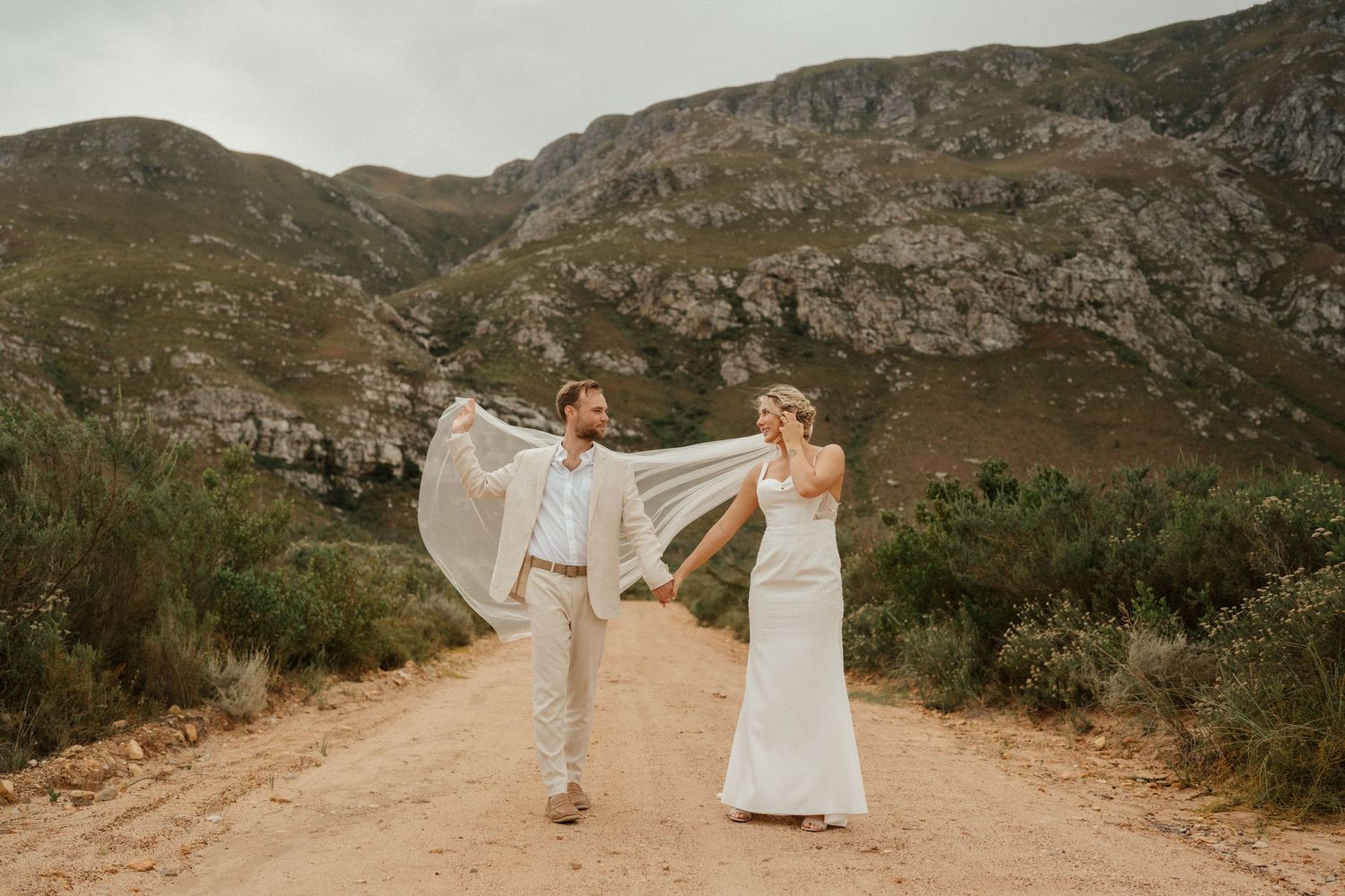 A romantic wedding couple walks down a dirt road with majestic mountains in the backdrop.