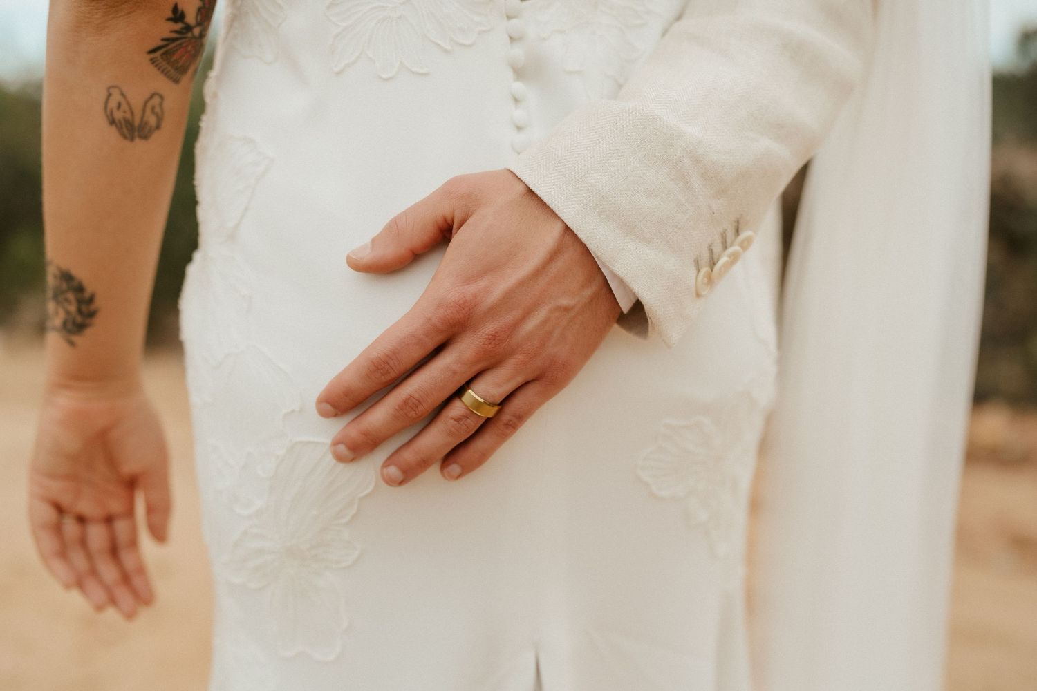 Close-up detail shot of a wedding ring and tattooed hand against white wedding attire.