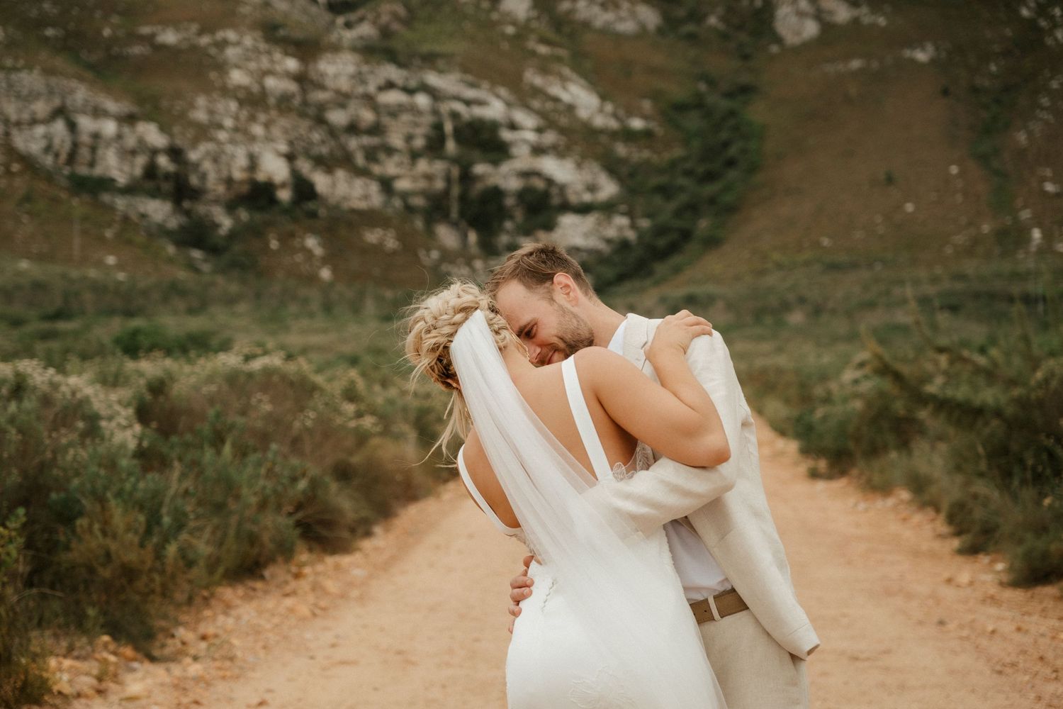 A candid moment between newlyweds embracing on a mountain trail with dramatic landscape views.