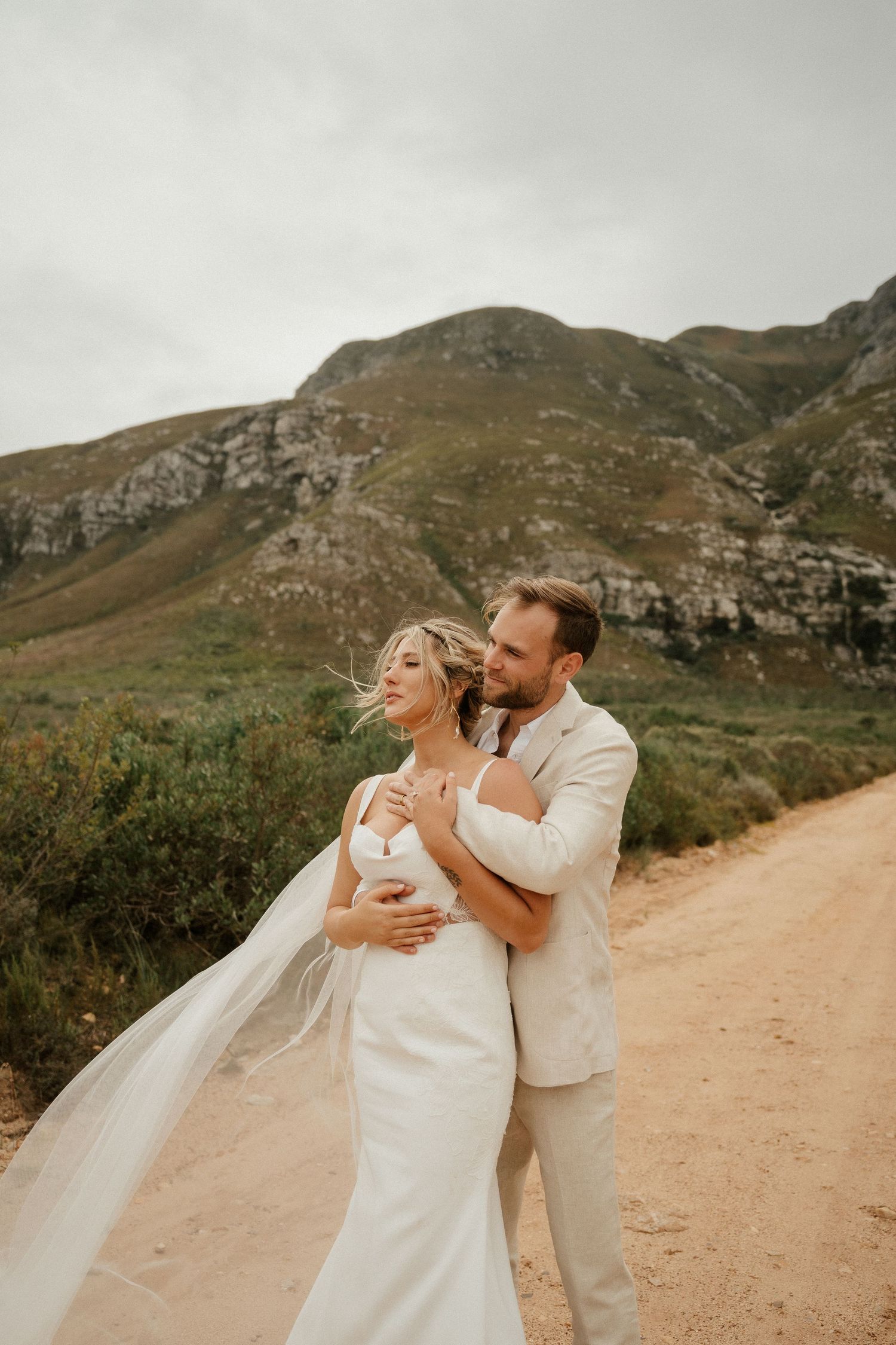 A romantic wedding photo of a couple embracing on a dirt path with majestic mountains in the background.