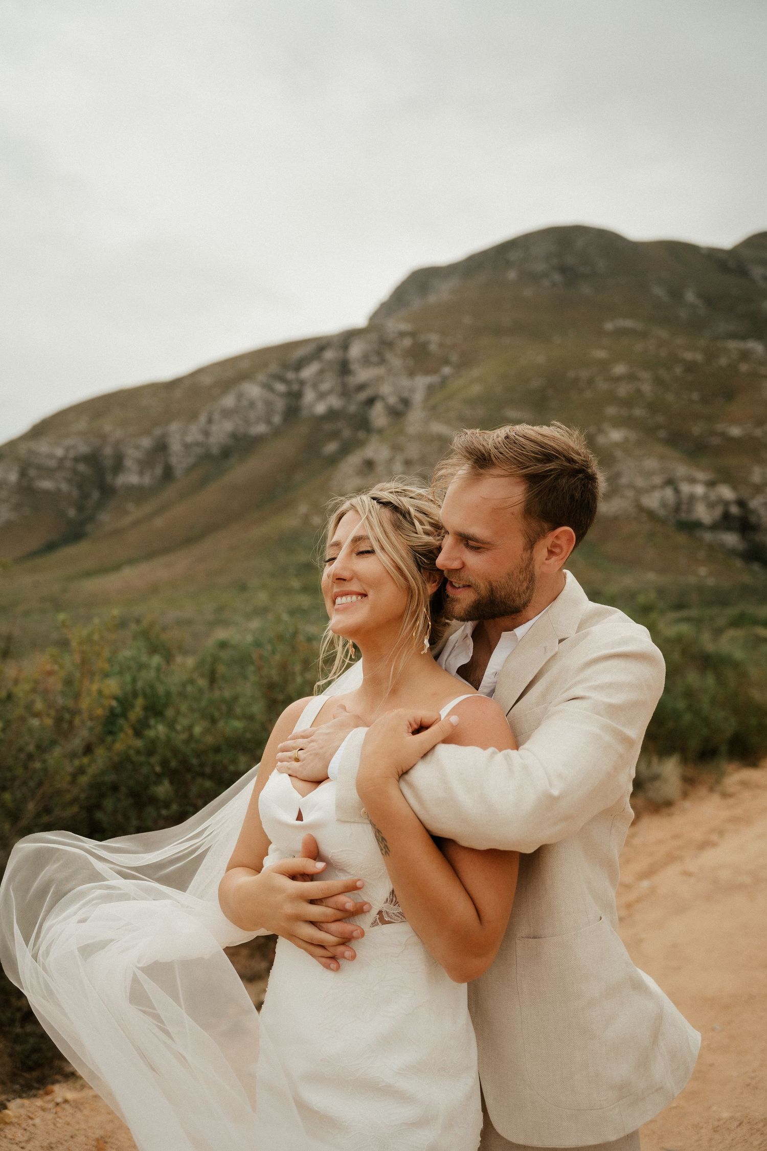 Joyful wedding embrace on a mountain trail with dramatic cliffs in the background.